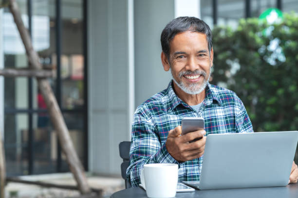 Smiling happy mature asian man with white stylish short beard using smartphone gadget serving internet at coffee shop cafe outdoor. Old indian or hispanic man using network technology with confident.