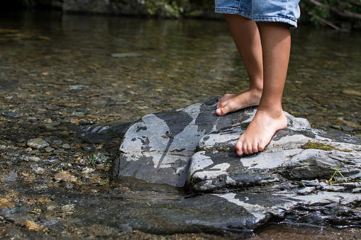 LEGS OF A CHILD PLAYING AND STANDING OVER A BIG STONE IN THE PEBBLES RIVER SHORE ON SUMMER