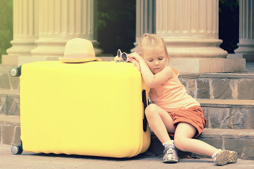 The little girl fell asleep sitting on the stairs. She leaned her elbows on the big yellow suitcase.