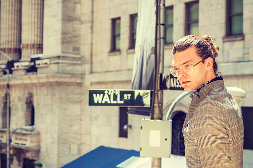 Portrait of Young Hispanic American Male College Student in New York City, with hair bun, wearing glasses, dressing in brown patterned jacket, standing on Wall Street, thinking, lost in thought.