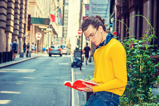 Young Hispanic American Man with hair bun, wearing glasses, yellow long sleeve T shirt, small black scarf around neck, sitting by green plants on old street in Manhattan, New York. reading red book.\