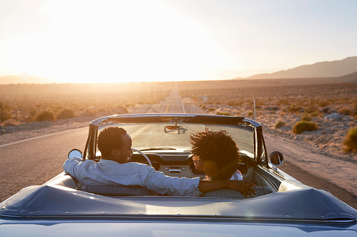 Vista posterior de la pareja en viaje por carretera conduciendo el coche descapotable clásico hacia la puesta del sol photo