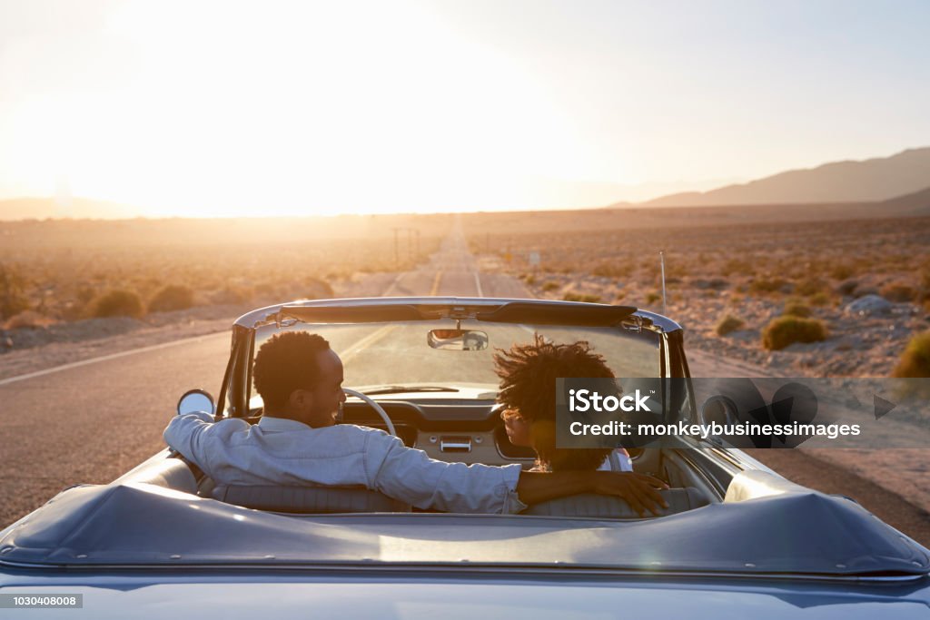 Vista posterior de la pareja en viaje por carretera conduciendo el coche descapotable clásico hacia la puesta del sol - Foto de stock de Coche libre de derechos
