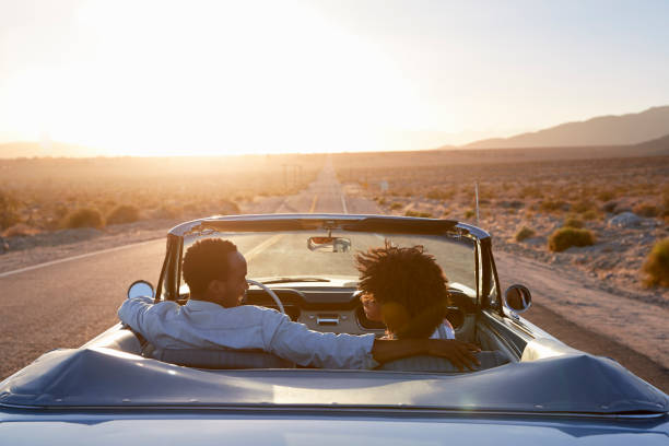 vue de l’arrière du couple en voyage sur la route conduisant une voiture décapotable classique vers le coucher du soleil - aventure photos photos et images de collection