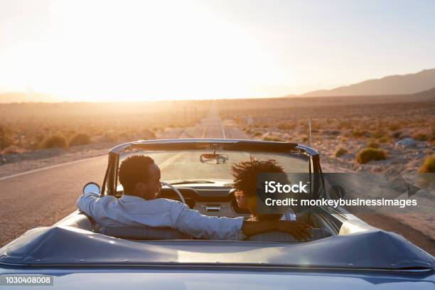 Rear View Of Couple On Road Trip Driving Classic Convertible Car Towards Sunset Stock Photo - Download Image Now