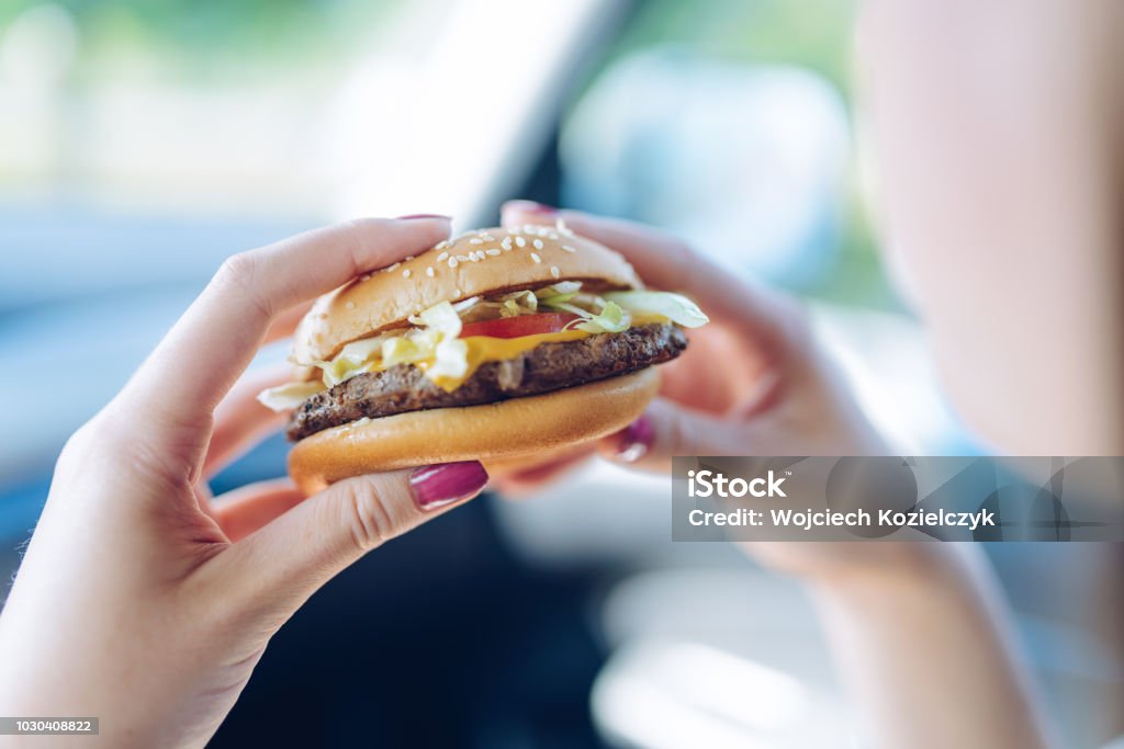 Ragazza che tiene un hamburger tra le mani seduta in macchina. Concetto alimentare malsano - Foto stock royalty-free di Fast Food - Ristorante