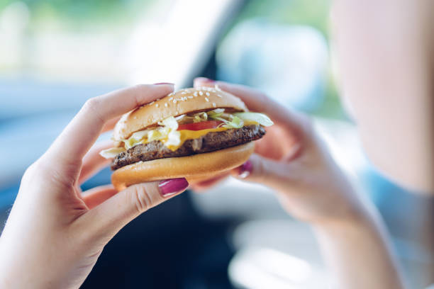 menina segurando um hambúrguer em suas mãos, sentado em um carro. conceito de alimentação saudável - restaurante de fast food - fotografias e filmes do acervo