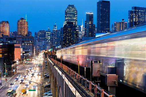 Train approaching  elevated subway station in Queens at dusk, New York. Financial buildings and New York skyline are seen in the background, on the left below can be seen a busy street full of cars at rush hour, USA.