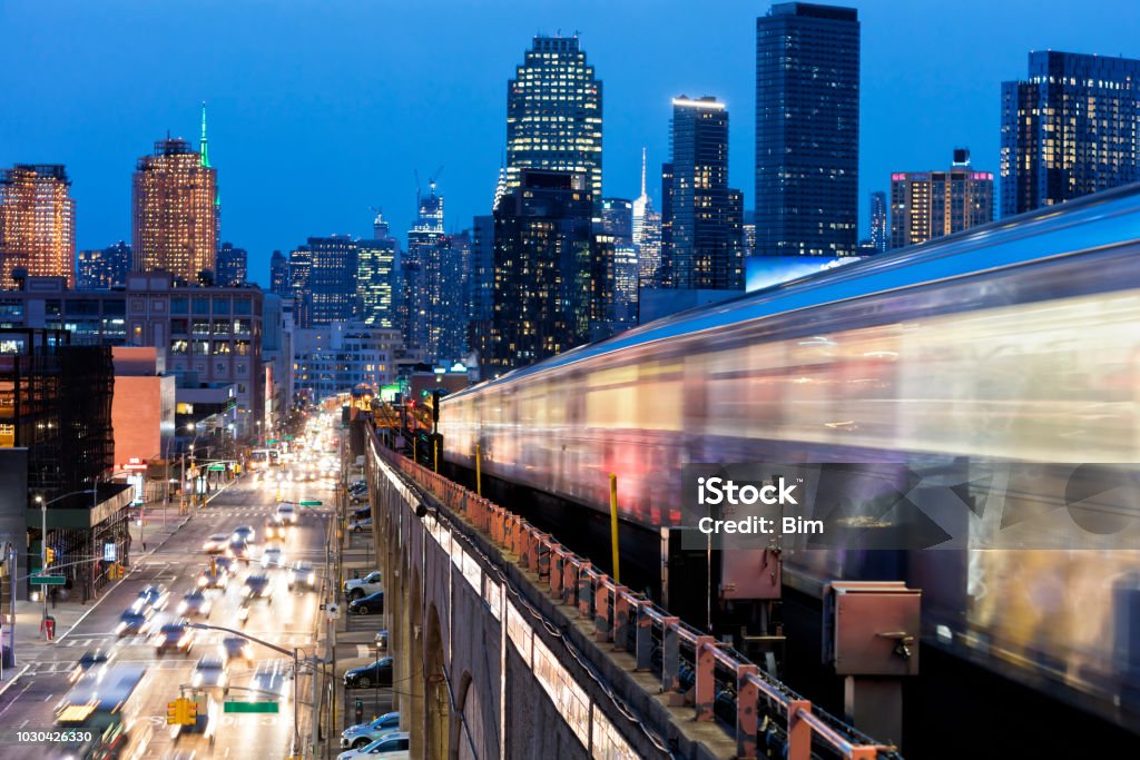 Subway Train Approaching  Elevated Subway Station in Queens, New York Train approaching  elevated subway station in Queens at dusk, New York. Financial buildings and New York skyline are seen in the background, on the left below can be seen a busy street full of cars at rush hour, USA. New York City Stock Photo