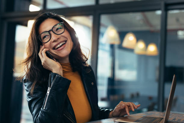 Smiling business woman in casuals talking on phone Cheerful asian female talking on mobile phone while sitting on desk with laptop. Business woman in casuals making a phone call and laughing. woman lifestyle stock pictures, royalty-free photos & images