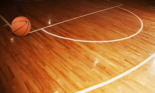 A shot of a female teenage athlete sitting on the floor during half time of a basketball game. She is smiling and looking into the camera. She is socialising with her team mates with a basketball by her side.