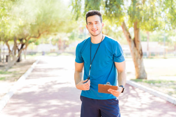 Young running coach holding clipboard and whistle while standing on footpath in park
