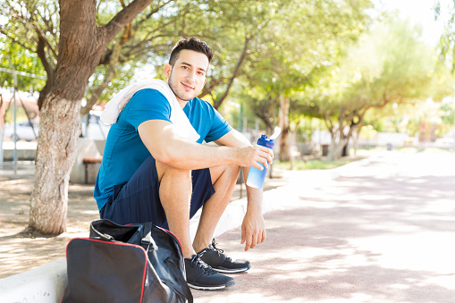 Tired male runner holding waterbottle while sitting on footpath in park