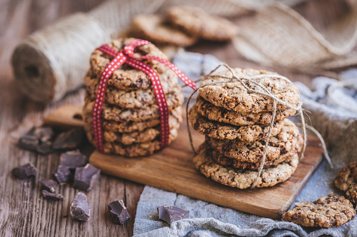 Homemade chocolate chip cookies. The cookies are  tied together with ribbons, one of the piles with a red and white ribbon. The cookies are on a wooden chopping board and there are pieces of chocolate next to it.