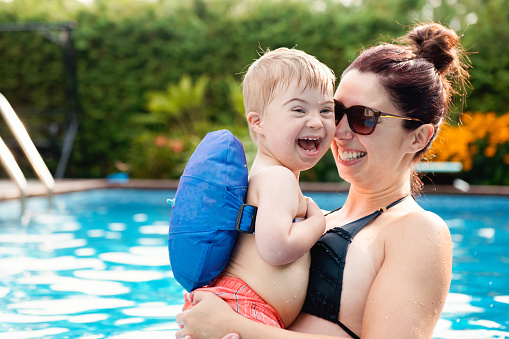 Three years old Little boy with Down syndrome having fun in the swimming pool with his family.  His mother holds him in her arms. Color and horizontal Photo was taken in Quebec Canada.