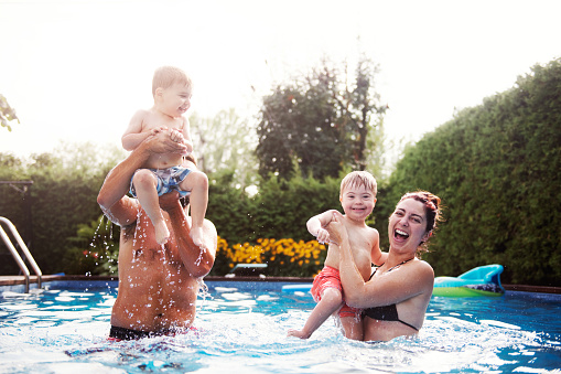 Three years old Little boy with Down syndrome having fun in the swimming pool with his family. His uncle holds him in his arms. His mother holds her two-year-old brother in her arms. Color and horizontal Photo was taken in Quebec Canada.