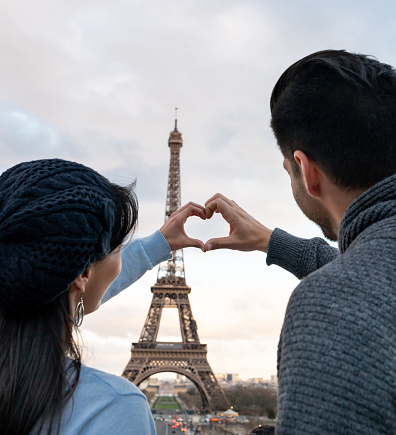 Portrait of a loving couple on a romantic getaway in Paris making a heart shape with their hands in front of the Eiffel Tower - travel concepts