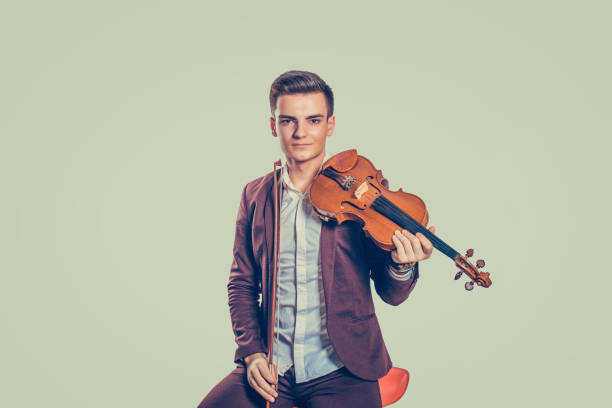 Young handsome man in elegant jacket sitting on chair with classic violin looking at camera