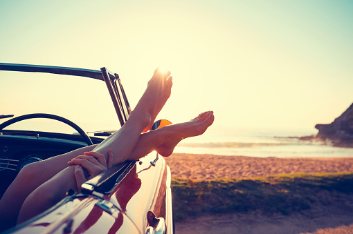 Convertible Car with womans feet hanging out of the window. The car is parked at the beach at sunrise. Very relaxing vacation or road trip image. Car is a convertible. copy space