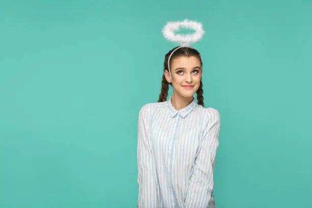 Photo of Happy funny girl in striped blue shirt and pigtail hairstyle, standing with halo on her head and looking away with smile and dreaming face