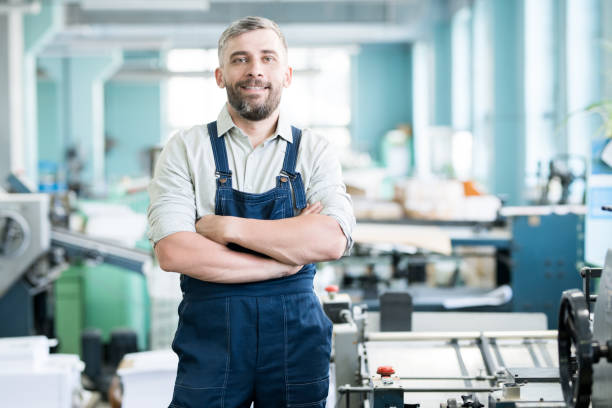 istock Cheerful content confident bearded typography worker in overall crossing arms on chest and looking at camera while standing among printing machines 1030642108