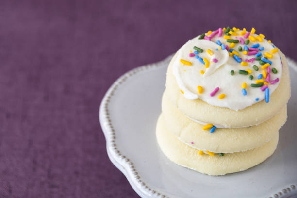 Sugar Cookies Three frosted sugar cookies, white frosting, colored sprinkles stacked on a white cake plate on a purple background white sugar cookie stock pictures, royalty-free photos & images