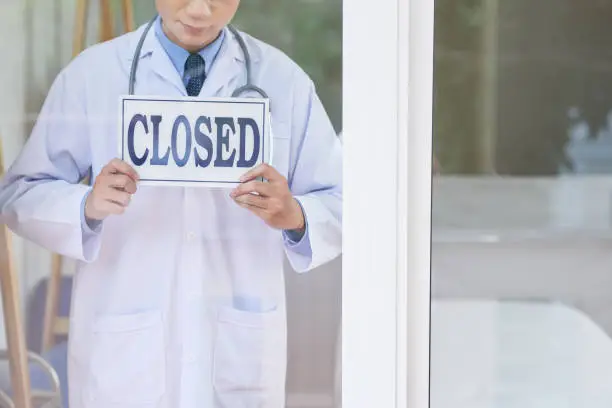Anonymous guy in medical uniform holding sign with closed writing while standing behind glass in office