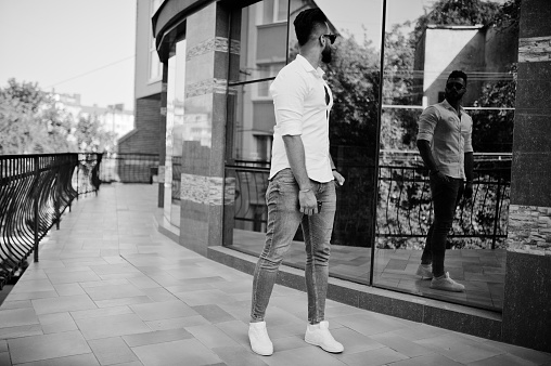 Stylish tall arabian man model in white shirt, jeans and sunglasses posed at street of city. Beard attractive arab guy against modern building.