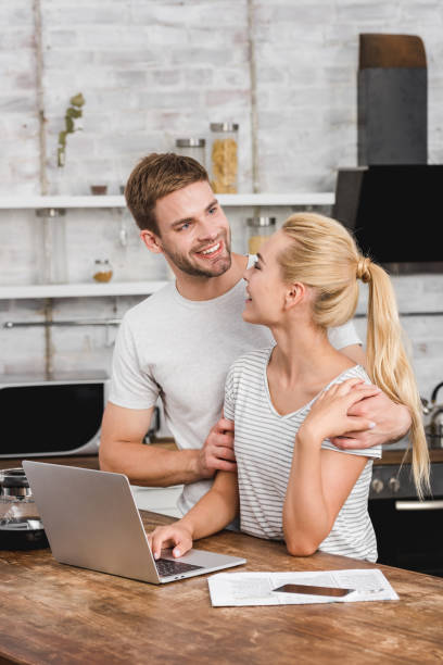 istock smiling boyfriend hugging girlfriend in kitchen while she working with laptop 1030783590
