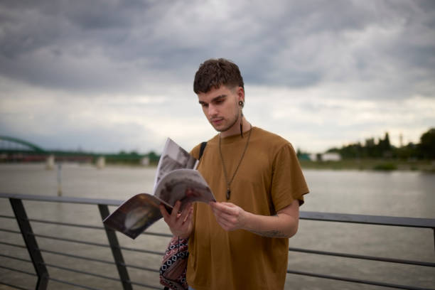 Portrait of young gay man somewhere in the city near the river while reading a magazine.