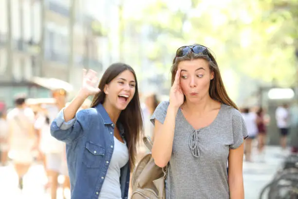 Happy teen greeting waving hand and friend ignoring her in the street