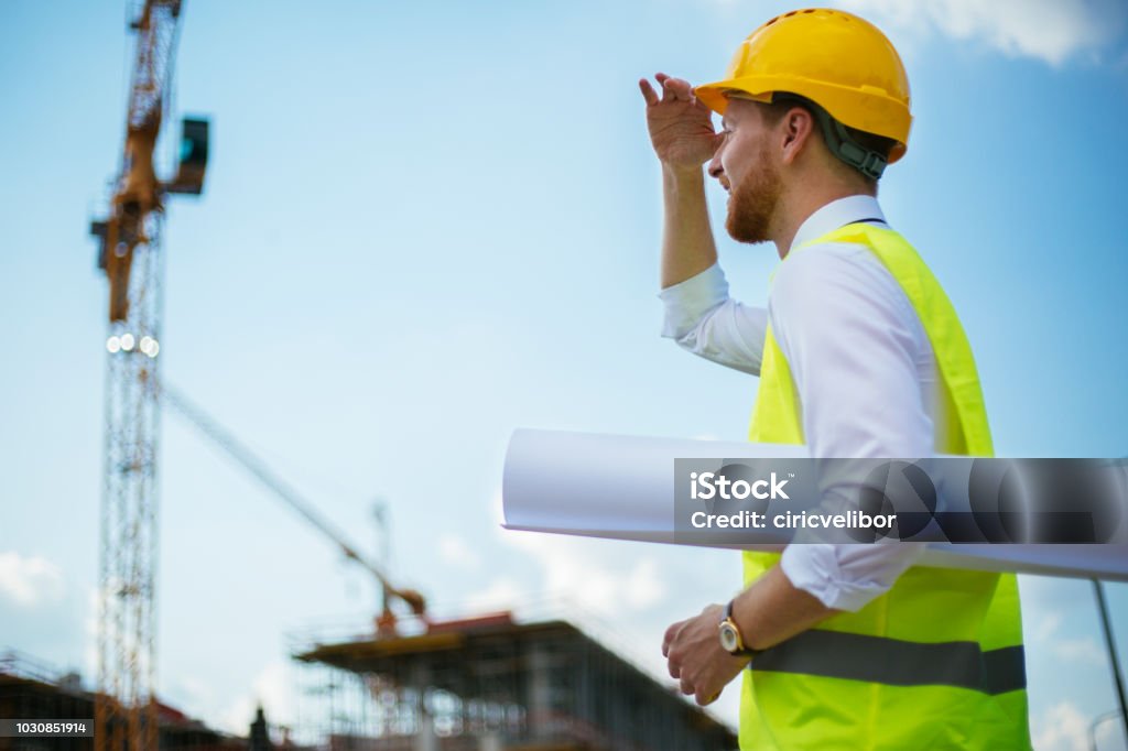 Young architecte looking at works on construction site Young construction worker with blueprints, yelow helmet, checklist on construction site Adult Stock Photo