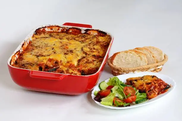 Baked layers of aubergine, cheese and tomato topped with melted cheese served with salad and bread with the remainder of the baked aubergine in a red ceramic baking dish to the rear.