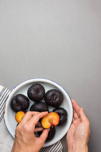 Young caucasian woman puts halved dark red purple plums with bright yellow reddish flesh on white plate. Rustic Style. Autumn harvest. Making preserves