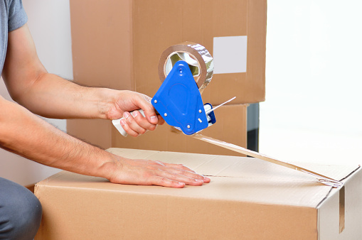 Shot of an unidentifiable young man closing a cardboard box with tape at home