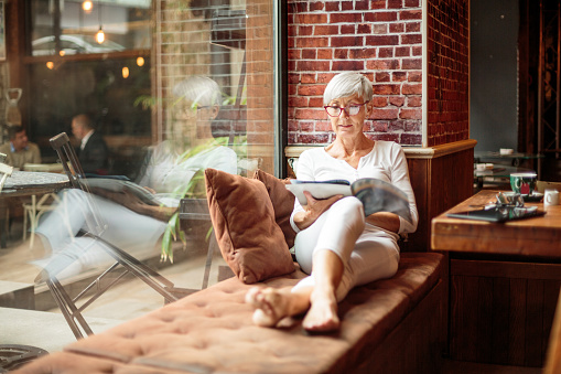 Portrait of active senior woman with eyeglasses in coffee shop while reading a magazine.