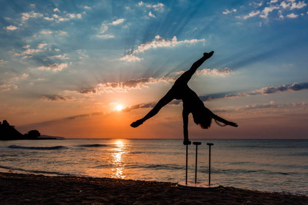 Flexible Circus Performer, silhouette of a female acrobat practicing on the beach by the sea at sunrise, with the sun rising from the horizon Beautiful photography of a girl acrobat practicing on the beach by the sea at sunrise, with the sun rising from the horizon. The flexible circus performer is doing her morning exercise on the sandy beach, while the red sun at sunrise is rising from the edge of the water. The horizon of the Black Sea is awesome, the sun rays are all across the sky. The sun and the sunbeams are reflecting in the sea water surface, and small waves are hitting the sand. The female practicing acrobatics is a silhouette She stretches her legs and holds them upside down, making handstand, in perfect balance. The dusk is a perfect time for training gymnastics and the clouds and landscape are sensational. The photo is taken in August, in summer, just before coming of autumn, near the Old Town of Nessebar, in the Burgas area, in Bulgaria. The sunrise looks like a sunset. acrobatic gymnastics stock pictures, royalty-free photos & images