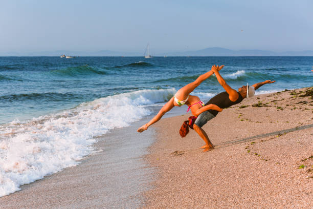 Beautiful photography of two acrobats practicing on the beach by the sea in the morning, at sunrise, with the sea horizon in the background. The flexible circus performers, one male and one female, are doing their morning exercise on the sandy beach. The horizon of the Black Sea is awesome, the sky is blue and clear, with scattered clouds. Waves are hitting the sand. The man and woman practicing acrobatics are a silhouette. She stretches her legs and holds them upside down, making handstand, in perfect balance. The man is showing strength, holding the female acrobat steady. The dawn and the dusk are the perfect time for training gymnastics and the clouds and landscape are sensational. The photo is taken in August, in summer, just before coming of autumn, near the Old Town of Nessebar, in the Burgas area, in Bulgaria. The sea is blue.