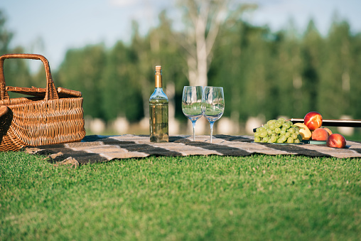 picnic with glasses, bottle of white wine, fruits and wicker basket on blanket on grass