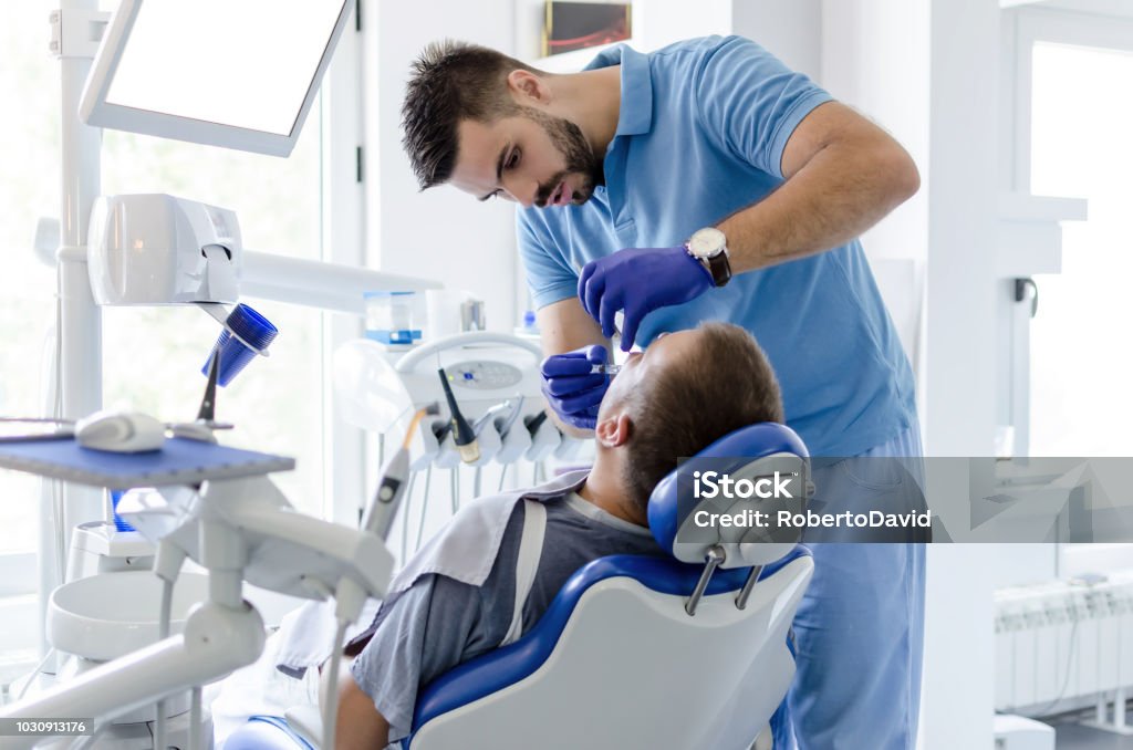 Dentis giving anesthesia to patient Image of male dentist in uniform giving anesthesia to male patient in stomatology office. Dentist Stock Photo