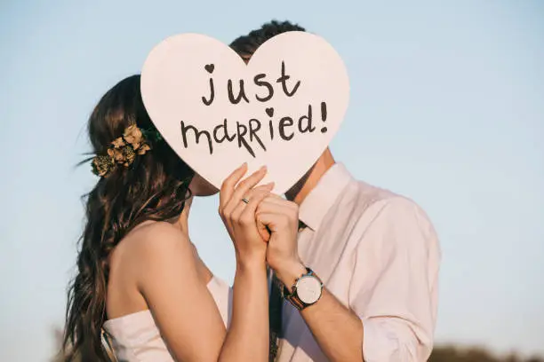 Photo of young wedding couple kissing and holding heart with just married inscription