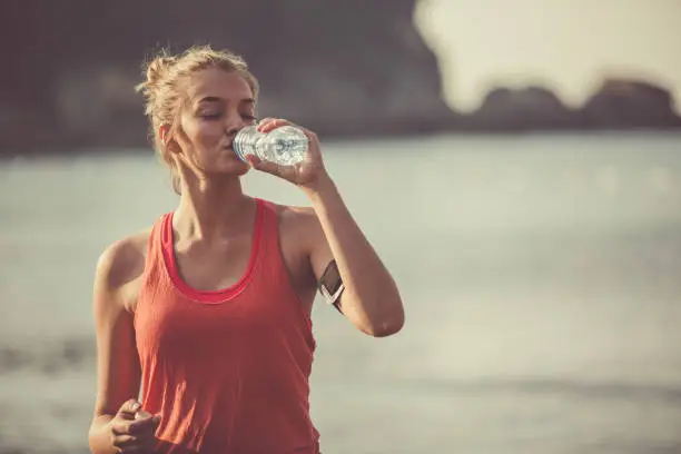 Beautiful young woman is drinking water from a bottle during her morning training outdoors. She is doing exercises at the beach.