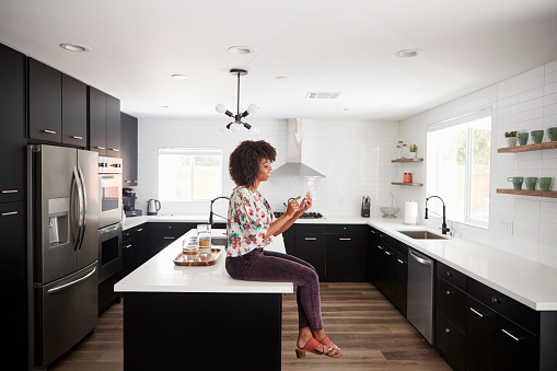 Woman At Home Sitting On Kitchen Island Whilst Using Mobile Phone