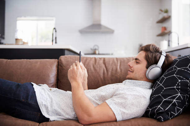 homme portant des casques sans fil couché sur le canapé à la maison en streaming depuis mobile - lying on side audio photos et images de collection