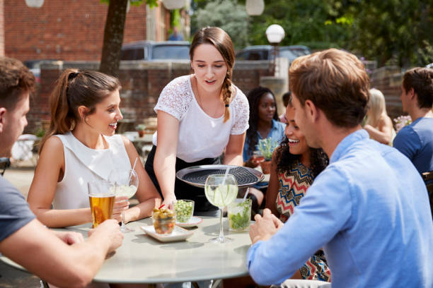 cameriera che serve bevande a un gruppo di amici seduti a tavola nel giardino del pub godendosi un drink insieme - giardino di birra foto e immagini stock