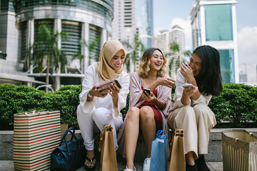 Multi-ethnic group of women sitting in front of the Petronas towers, using their smart phones, Kuala Lumpur, Malaysia
