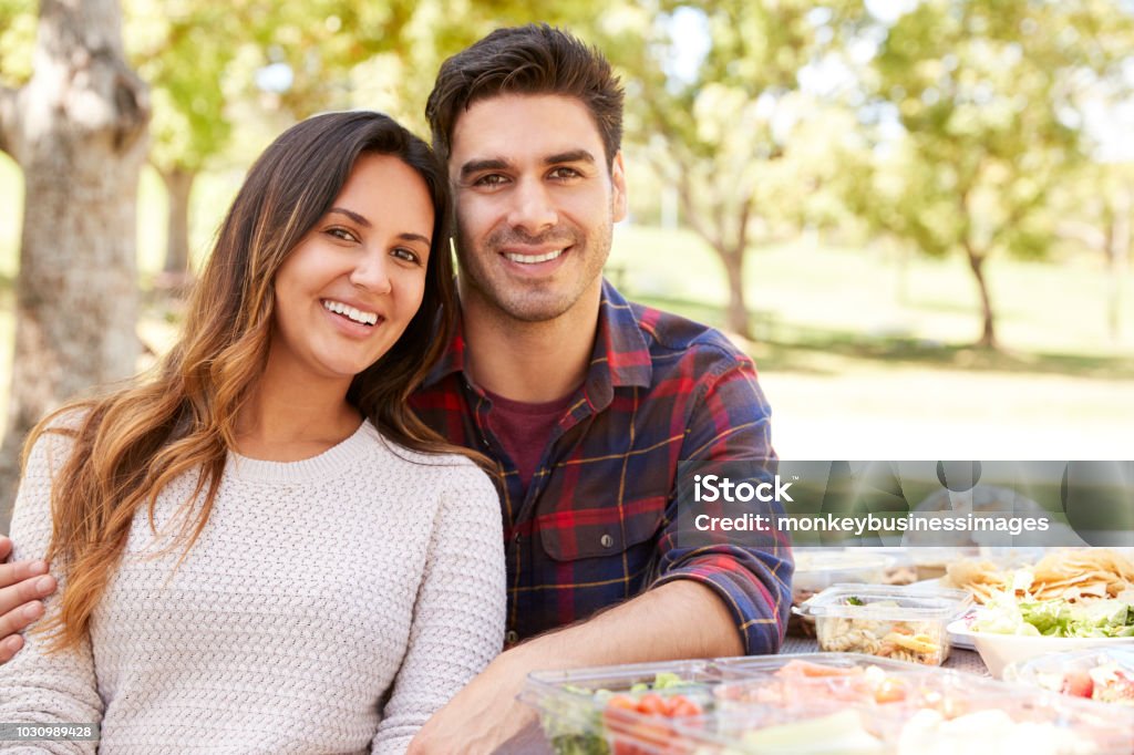 Jeune couple assis à la table de pique-nique, souriant à la caméra - Photo de Tous types de couple libre de droits