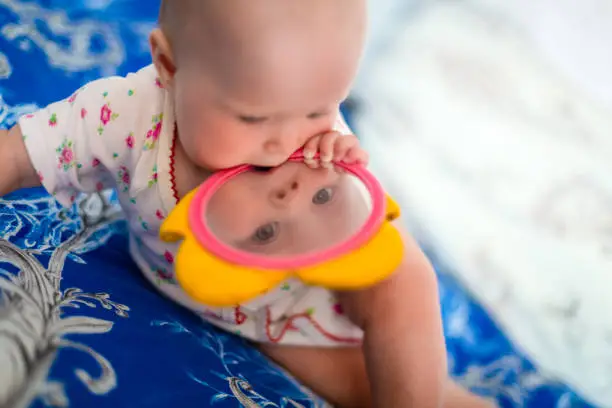 Photo of Adorable and cute baby girl sits on the bed and plays with toys that is children mirror. Small child is holding flower toy in the mouth and looking through mirror.