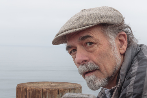 Close up of thoughtful, serious senior man with a gray beard wearing a tweed cap looking at camera with ocean horizon in background