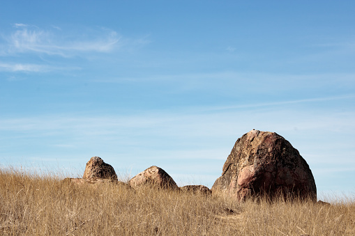 One large boulder and three smaller big rocks set against blue sky with white wispy clouds with dry grass in foreground